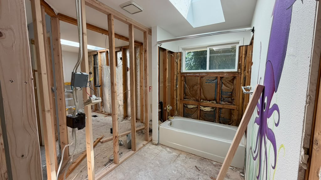 Gutted split-level bathroom showing the original fiberglass tub and remnant wall mural before reconfiguration