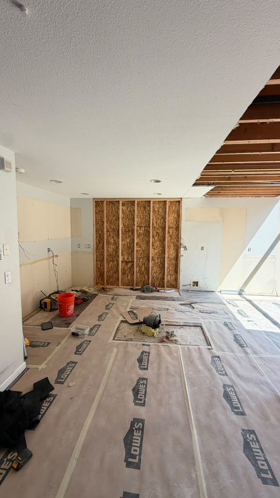 A residential kitchen undergoing renovation with protective paper flooring laid across the space, bare drywall, exposed wooden ceiling beams, and construction materials including a red bucket and tools scattered throughout. The room shows various stages of remodeling work with framed wall sections, electrical outlets being installed, and temporary plastic sheeting covering work areas. Recessed lighting fixtures are visible in the ceiling, indicating ongoing installation of electrical and finishing work.