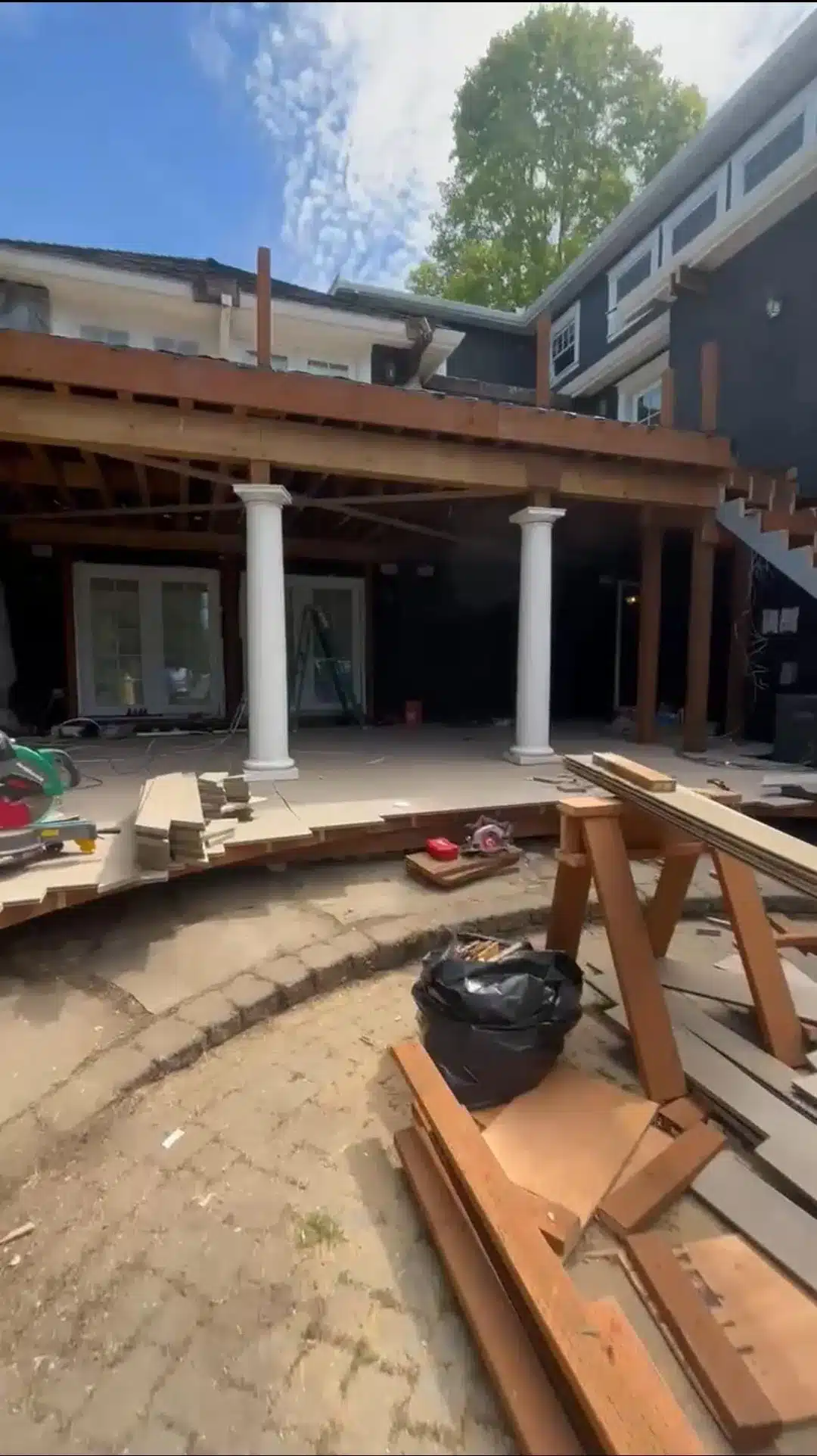 Patio under construction with exposed wood framing, two white columns, stacked lumber, and tools.