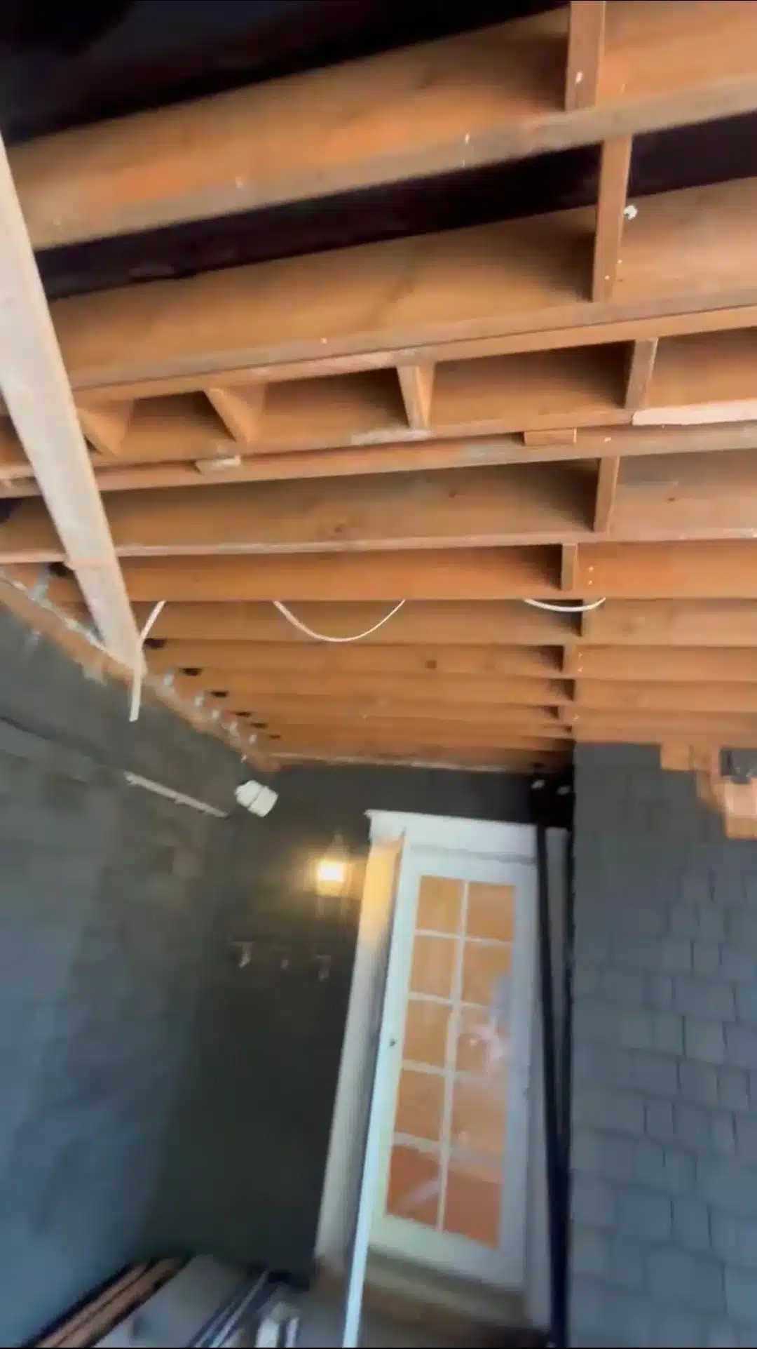 Exposed wooden ceiling joists with white wiring above dark shingle siding and a white-trimmed glass door.
