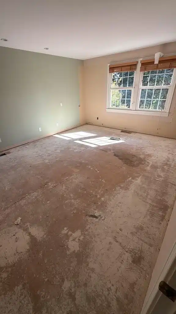 Empty room with green and beige walls, recessed lights, white-trimmed windows, and exposed plywood subfloor.